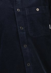 Granatowa koszula z korduroy z zapięciem na guziki, pięcioma czarnymi plastikowymi guzikami oraz kieszenią na piersi z detalem w postaci przeszycia.