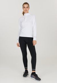 Weißes Langarm-Sportoberteil mit hohem Kragen und Halbreißverschluss, kombiniert mit schwarzen Leggings mit reflektierenden Akzenten und schwarzen Sportschuhen.