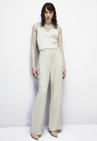 Camisón de satén beige superpuesto sobre una blusa larga de mangas transparentes y con lentejuelas; pantalones de pierna ancha con una textura suave; combinados con tacones en tono nude.