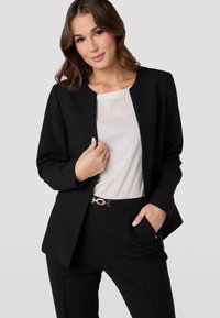 Černý blazer s pevnou strukturou, nošený přes bílé tričko, s předními kapsami a zlatým páskem, doplněný černými kalhotami.