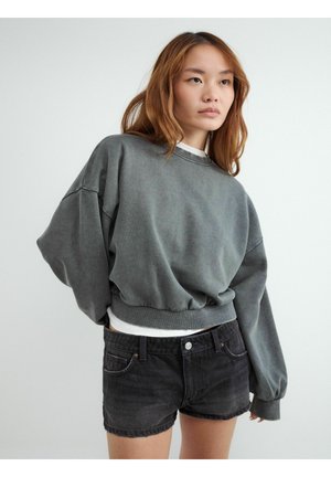 Młoda kobieta z długimi włosami ubrana w luźną szarą bluzę i czarne dżinsowe szorty, stojąca na jednolitym jasnym tle.