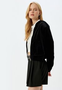 Felpa nera in velluto con zip, dotata di tasca frontale e orlo a costine, abbinata a una camicia bianca e pantaloni corti plissettati neri. Texture liscia.