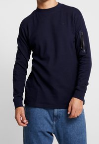 Marinblå långärmad skjorta i texturerad tyg med rund halsringning och en zip-försedd ficka på vänster ärm. Avslappnad passform med ribbade ärmslut.