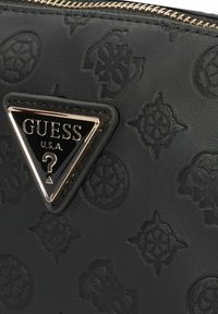 Zwarte leren portemonnee met reliëfpatronen, een gouden driehoekig logoplaatje met "GUESS U.S.A." en een vraagtekenembleem. Met ritsluiting.