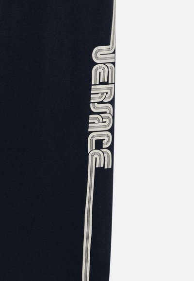 Námořnické tepláky s bílým vertikálním logem "Versace" na boku. Vyrobené z měkké, strukturované látky s kontrastními pruhy.