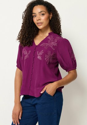 Mujer con cabello rizado, vestida con una blusa morada abotonada con patrones florales bordados y pantalones azules, de pie con una mano en el bolsillo.