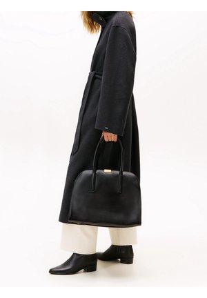 Personne portant un long manteau sombre et un pantalon clair, tenant un grand sac à main en cuir noir avec deux poignées, debout devant un fond blanc.