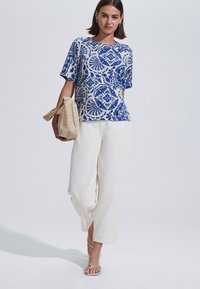 Sininen ja valkoinen kuviollinen lyhythihainen paita pyöreällä kaula-aukolla, yhdistettynä valkoisiin leveälahkeisiin housuihin ja beigenvärisiin sandaaleihin, kädessä kuvioitu laukku.