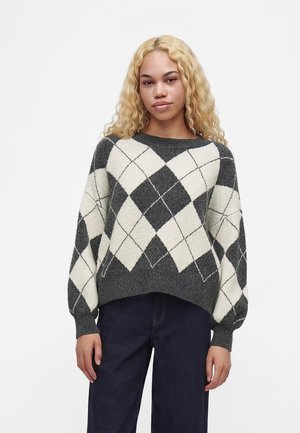 Jeune femme aux longs cheveux blonds bouclés portant un pull gris et blanc à motif losange et un pantalon foncé, debout devant un fond uni.