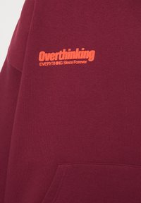 Bordó kapucnis pulóver narancssárga "Overthinking" felirattal a mellkason. Az anyaga puha, elején zsebével és sima textúrával rendelkezik.