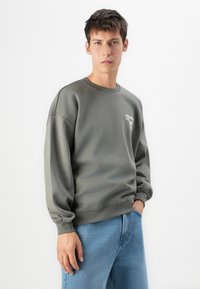 Young man wearing a gray sweatshirt with small white logo and light blue jeans, standing with one hand in pocket against a white background.