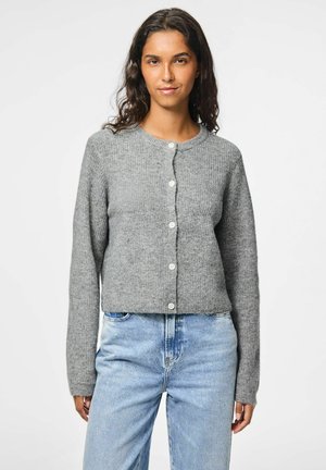 Grå kort cardigan i texturerad stickad tyg, med långa ärmar och fem vita knappar framtill. Kombinerad med ljusblåjeans.