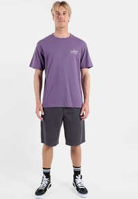 T-shirt violet en coton avec col rond et manches courtes. Shorts noirs, baskets montantes noires avec des accents blancs, et chaussettes rayées.