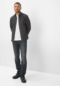 Camicia nera a fantasia con bottoni sopra una t-shirt bianca, abbinata a jeans scuri e scarpe nere stringate. La camicia presenta una stampa geometrica.