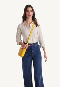 Sac à bandoulière jaune de forme rectangulaire, texture lisse et bandoulière ajustable, associé à une chemise légère et un pantalon large.