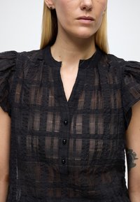 Svart, teksturert bluse med puffete korte ermer, med bølget krage, knapplukking foran og et rutenettmønster med subtil glans.