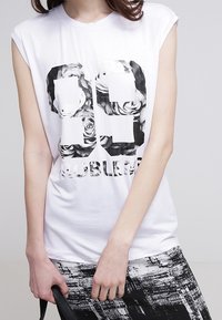 T-shirt bianca senza maniche con testo nero audace stampato "99 PROBLEMS", abbinata a pantaloni neri e bianchi con fantasia. Tessuto morbido, vestibilità casual.