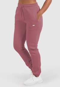 Rosa Sweatpants aus weichem Stoff mit elastischem Bund, seitlichen Taschen und einem kleinen Logo an der Hüfte. Kombiniert mit weißen Sneakern.