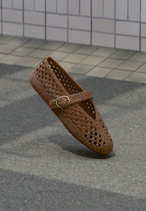 Bruine gevlochten platte schoen met een ronde neus, geperforeerd ontwerp en decoratieve gouden gesp, rustend op een textuurachtige ondergrond.