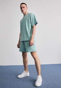 Set de tricou și pantaloni scurți din bumbac de culoare verde mentă. Croială lejeră, mâneci scurte, detaliu cu logo pe piept. Adidași albi, stând pe un covor albastru.