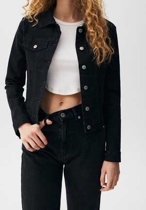 Personne aux cheveux bouclés portant une veste en jean noire et un pantalon avec un top court blanc, tenant la ceinture de son pantalon.