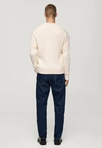 Cream cable-knit sweater with ribbed hem, paired with dark blue jeans. The back view shows a fitted silhouette and minimal detailing.