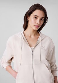 Sweat à capuche zippé beige clair en tissu lisse, avec une capuche à cordon, une poche kangourou et un logo discret sur la poitrine.