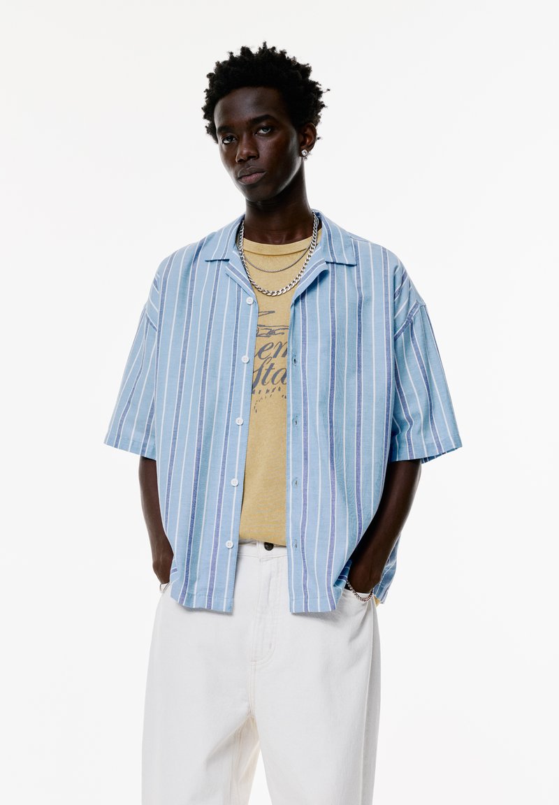Camicia a maniche corte blu chiaro con righe verticali, abbinata a una t-shirt grafica beige e pantaloni bianchi. Presenta bottoni e una vestibilità comoda.