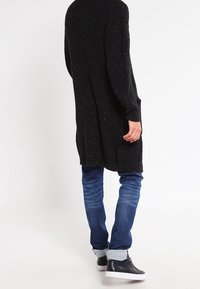 Černý texturovaný pletený cardigan, dlouhé rukávy, volný střih, do kolen, nošený přes modré džíny a černé tenisky.