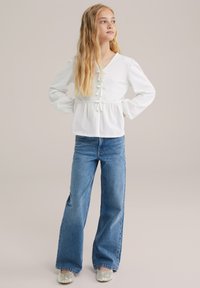 Vit texturerad långärmad topp med V-ringning, knappar framtill och banddetaljer, ihop med blå vida jeans och glittriga skottar.