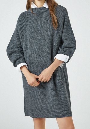Osoba ubrana w luźną, prążkowaną, ciemnoszarą sukienkę swetrową z widocznymi białymi mankietami i kołnierzykiem koszuli, z dłońmi złożonymi przed sobą.