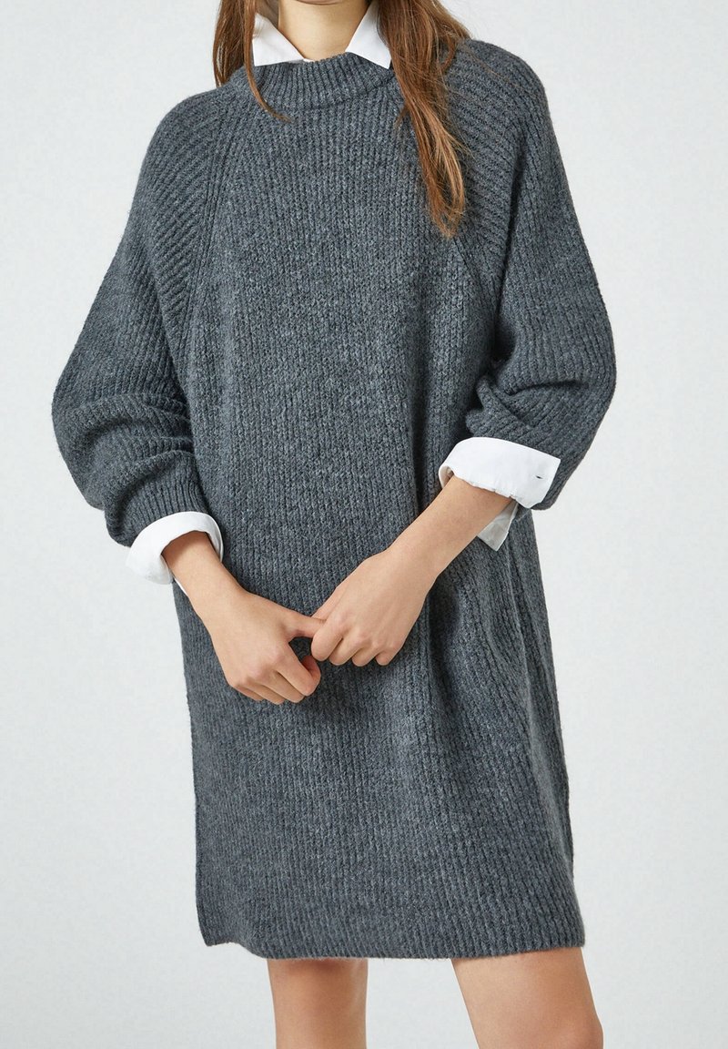 Personne portant une robe pull ample en maille côtelée gris foncé, avec les poignets et le col de chemise blancs visibles, mains jointes devant.
