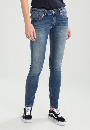 Personne portant un jean bleu slim et des baskets noires et blanches, debout avec les bras détendus le long du corps.