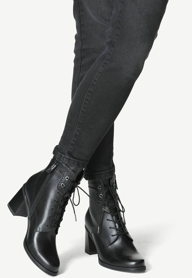 Bottes à la cheville en cuir noir avec talons carrés, lacets sur le devant, fermetures éclair latérales, portées avec un jean foncé ajusté.