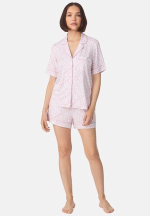 Femme aux cheveux courts bruns portant un haut de pyjama à manches courtes à motif rose clair et un short assorti, debout pieds nus sur fond blanc.
