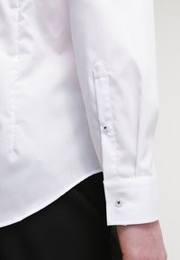 Camicia bianca, in tessuto di cotone, maniche lunghe con polsini a bottone, colletto a punta e orlo curvo, con piccoli bottoni argentati.