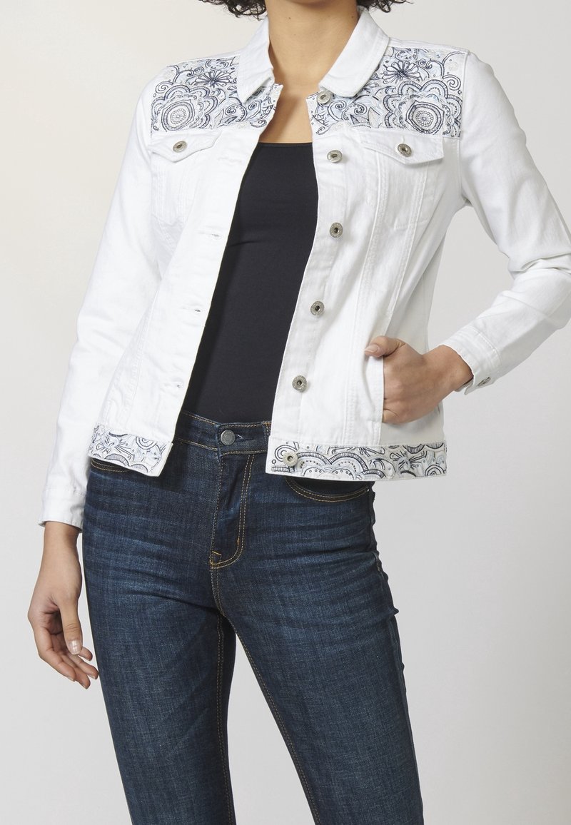 White denim jacket featuring a patterned embroidery on the chest and cuffs. The design includes blue accents and a classic collar.
