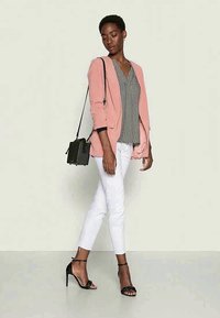 Blazer rosa claro sobre uma blusa estampada, calças brancas slim-fit e saltos pretos com tiras. Inclui uma pequena bolsa preta de mão.