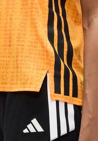 Πορτοκαλί αθλητικό τοπ με υφασμένο μοτίβο, μαύρες κάθετες ρίγες και πλευρικές σχισμές, σε συνδυασμό με μαύρο σορτς με λευκές λεπτομέρειες.