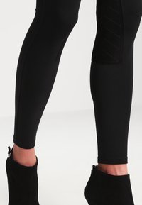 Nohy v černých přiléhavých kalhotách s prošívanými kolenními záplatami a černých kotníkových botách se bočními zipy.