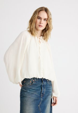 Žena s vlnitými blond vlasmi, oblečená v voľnej bielej blúzke s balónovými rukávmi a modrej rifľovej sukni na jednoduchom pozadí.