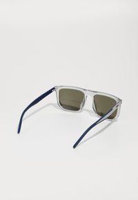 Ochelari de soare cu ramă din plastic transparent și lentile de culoare verde închis. Bratele sunt de culoare albastru marin, cu o textură netedă și un detaliu subtil de branding.