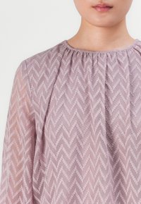 Light pink blouse with a zigzag pattern, made of sheer fabric, featuring a gathered round neckline and long sleeves. Textured details prominent.
