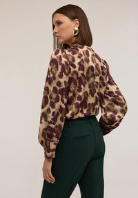 Blusa di satin con una stampa astratta di leopardo nei colori oro, viola e verde. Presenta maniche a palloncino e un polsino con bottoni, abbinata a pantaloni tailored verdi scuri.