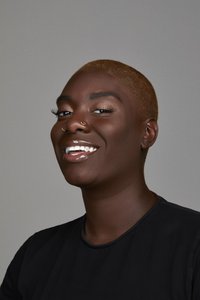 Portrait d'une personne avec une coiffure blonde très courte, une peau foncée, portant une chemise noire, avec plusieurs boucles d'oreilles et un anneau au nez.