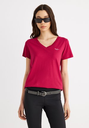 Γυναίκα με σκουρόχρωμα γυαλιά ηλίου, κόκκινη μπλούζα V-neck με λευκό λογότυπο, μαύρο παντελόνι και μαύρη ζώνη με καρφιά, σε απλό ανοιχτόφωτο φόντο.