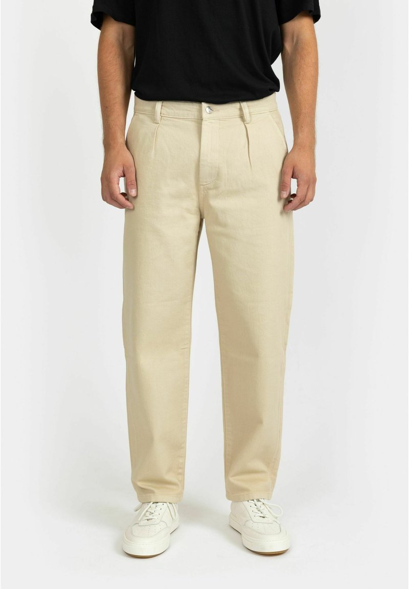 Pantaloni beige a pieghe realizzati in morbido tessuto misto cotone, con una vestibilità classica e due tasche laterali, abbinati a sneakers bianche.