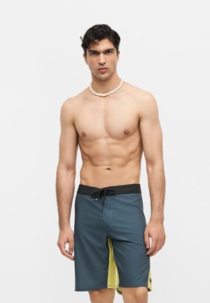 Homme torse nu portant un maillot de bain bleu et jaune et un collier de coquillages blanc, debout devant un fond clair uni.