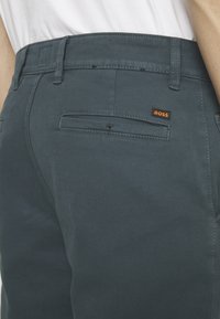 Pantalon bleu sarcelle foncé avec poche imperméable arrière et petite étiquette logo BOSS orange, porté par une personne en chemise blanche.