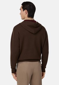 Hnedá mikina s dizajnom na chrbte, so šnúrkou na kapucni a rebrovanými manžetami, nosená cez béžové nohavice. Jemný materiál s voľným strihom.